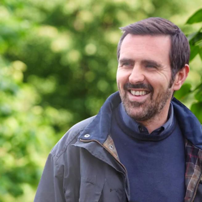 A smiling Adam Frost in navy knitwear and a coat casually looks off-camera in a luscious green garden. 