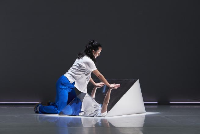 ID: A woman of Bundjalung and Ngāpuhi descent is pictured in a dimly lit room. Wearing a white shirt and bright blue trousers, she is knelt against the floor. The palm of her hands rest against a mirrored triangle as she leans her body forward into her reflection.