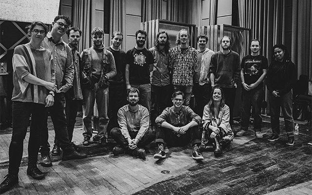 A black and white group shot of the Ancient Infinity Orchestra on a stage, featuring 15 members