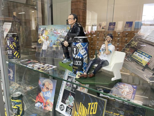 Display case featuring Leeds United Football Club memorabilia, including two figurines—one seated holding a bottle, the other standing beside a large "LOCO" can—set against a backdrop of celebratory fan photos, a "HERE WE GO WITH LEEDS UTD" sign, and club-themed magazines.