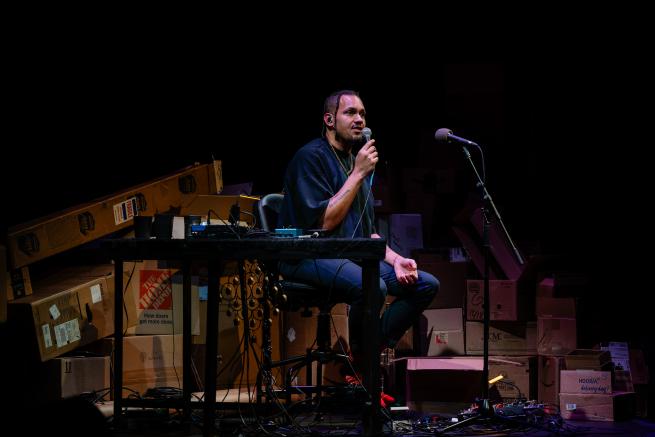 Ahamefule J. Olu sits on a stage surrounded by instruments. Wearing a black t-shirt and jeans, they are captured mid-performance, holding a microphone to their lips. Their hair is shaved at the sides with long cornrows on top, their face framed by their braids as they gaze forward. 