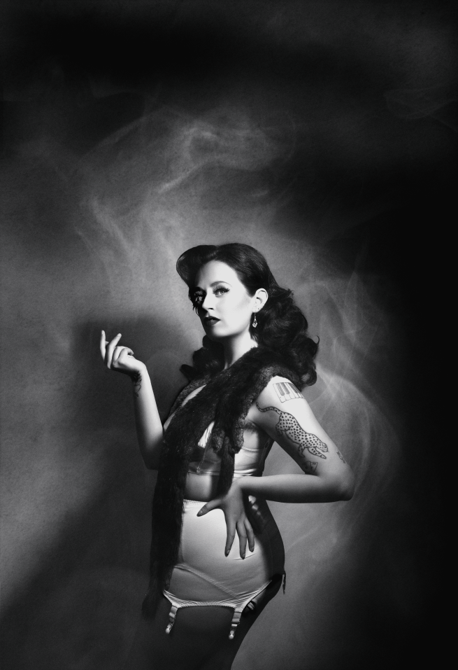 A black and white image of Selena Mersey,  a white woman with long brown hair, posing as the character Bisexuelle. She is wearing vintage-style lingerie and a stole, and her hair is waved. She has one hand on her hip and the other in the air as if holding a cigarette holder, and she is looking mysteriously into the distance.