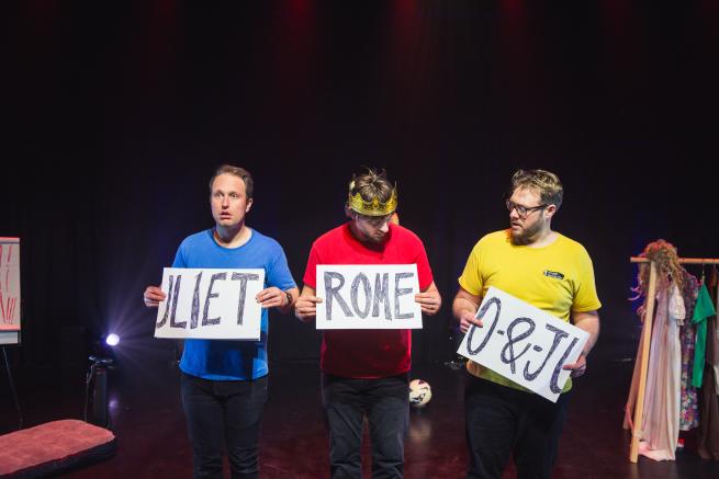 Three confused men on stage with signs reading Romeo and Juliet but in the wrong order. 
