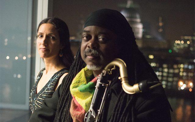 A man holding a saxophone, stood next to a woman with the US Capitol Building visible in the background through a window