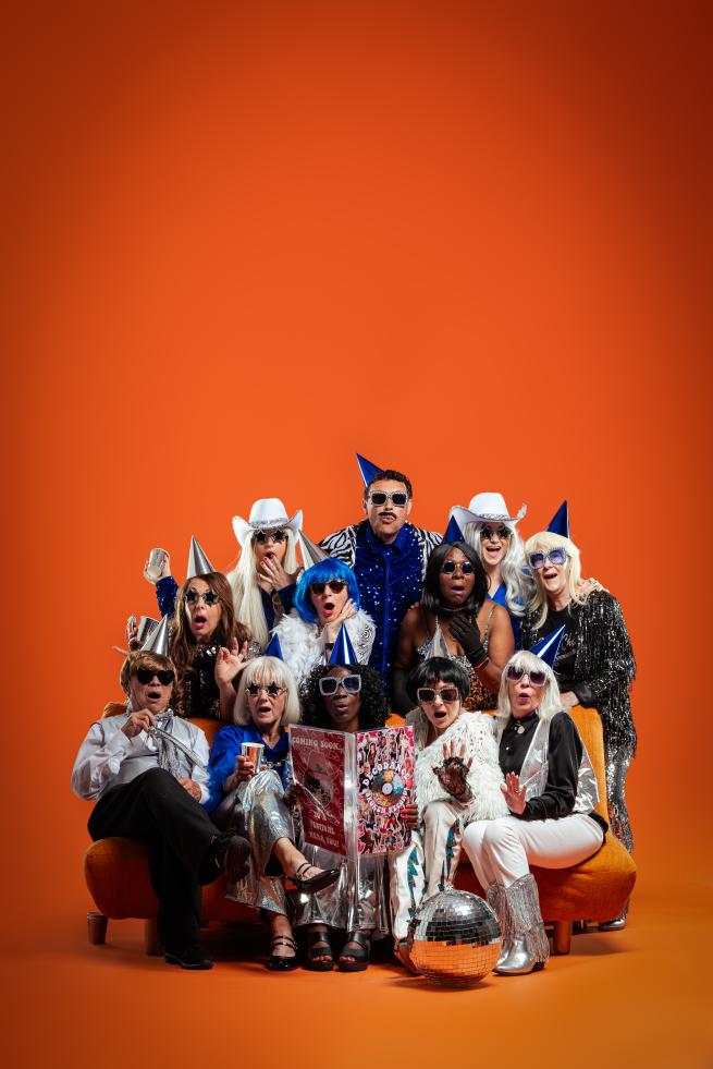 Performers looking shocked wearing party hats and disco costumes, reading a magazine.