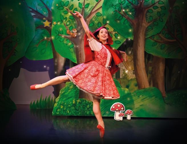 On a woodland background, a dancer wearing a red dress and a red cape balances on one foot.