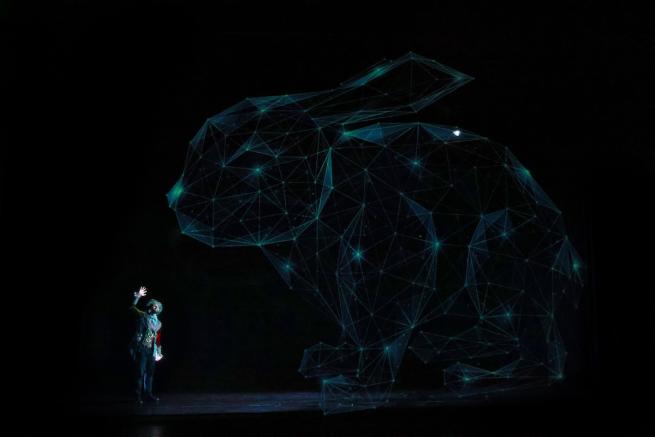 On a dark background, a dancer in a large top hat waves at a large bunny created with beams of light.