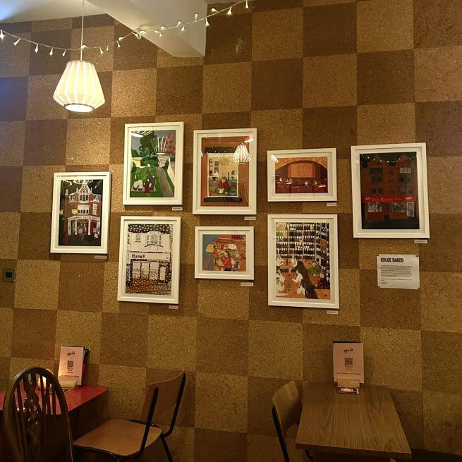 Ten gouache illustrations in white frames hung on a corkboard wall in a bar space. The room is dimly lit with fairy lights and candles. 