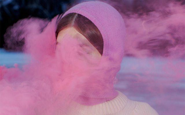 A person in a surreal scene wearing a pink headscarf, partially obscured by pink smoke