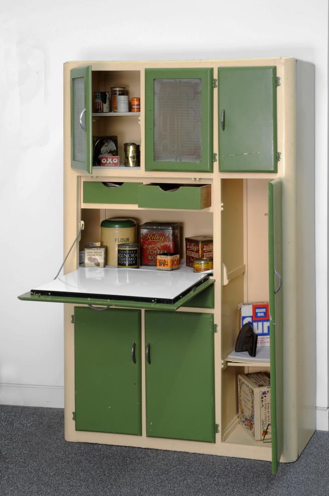 A picture of a mid 20th century kitchen unit with green doors and cream painted shelves, containing an array of tinned and dried foodstuffs.