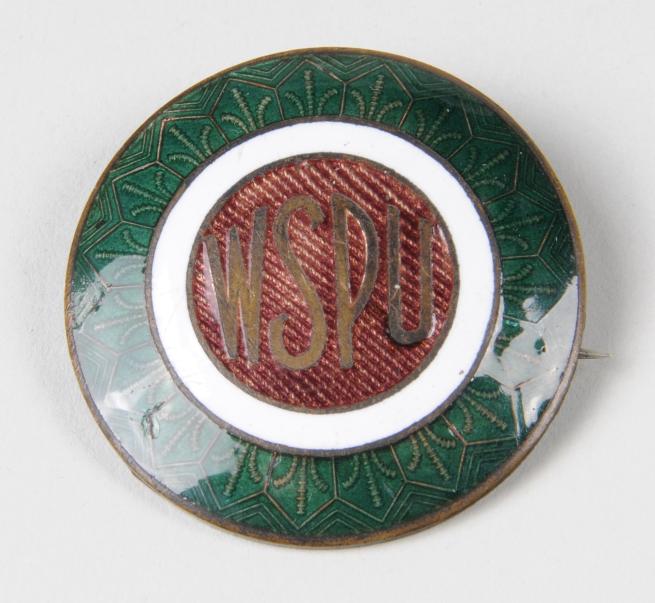 A circular pin badge bearing the inscription WSPU. Concentric green and white circles surround the central red circle which has gold lettering.