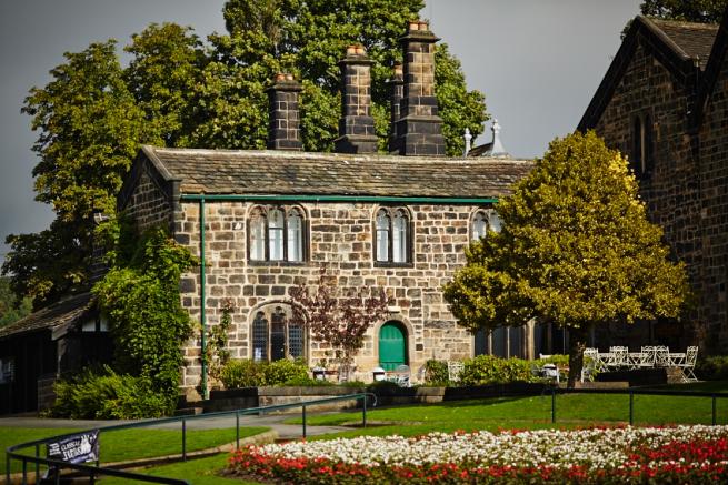 An image of a stone house on a hill. The window frames and doors are painted green. there are trees in the foreground and background and the sky is grey.