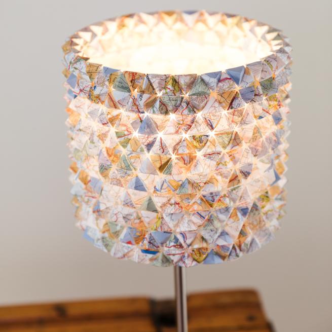 A close up photograph of a pastel coloured map lampshade on a wooden desk against a dark white background