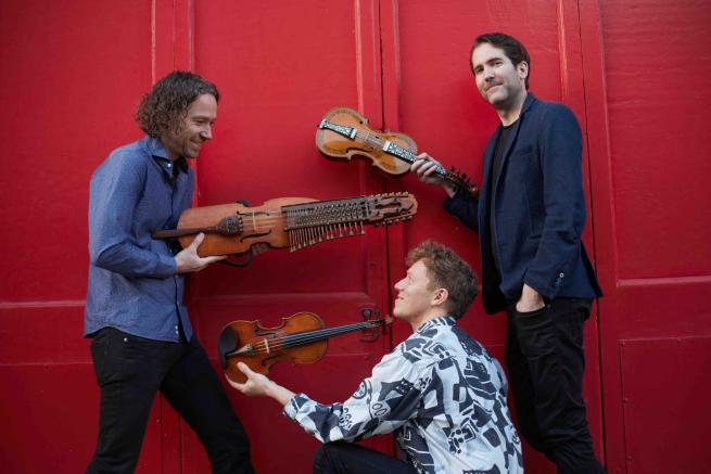 Three musicians holding their instruments up against a red wall. 