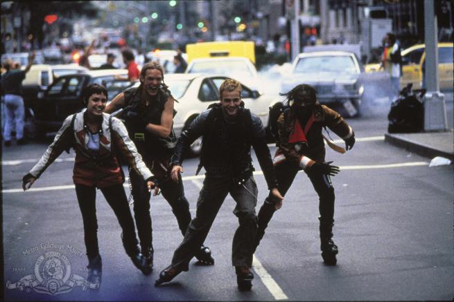 Four teenagers in cyberpunk outsits rollerblade down the middle of a busy street 