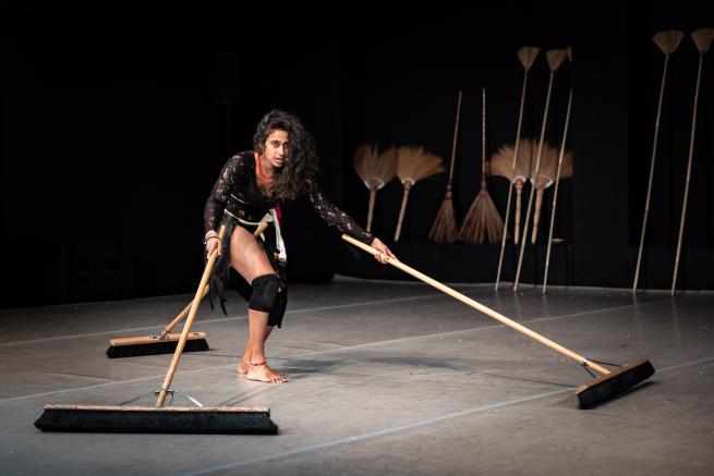 Venuri Perera, a young Sri Lankan woman dances under a spotlight. Her long brown curly hair cascades over the left side of her face as she stares into the distance. She is wearing a black lace leotard and black knee pads. Around her neck is a thick beaded necklace in red. As she hunches she pushes two brooms forward. The brooms are made out of wood and are long and wide. She has a broom tucked behind her, between her legs.