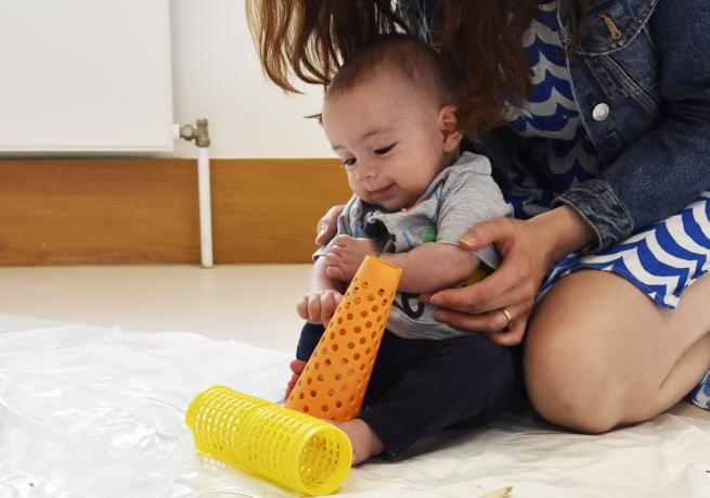 A baby sat on the floor, supported by its mother, playing with coloured plastic objects.