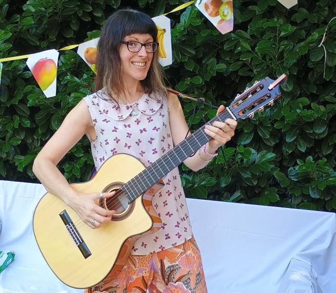 An image of a lady (Nicky Bray) with a guitar, smiling, in a garden with bunting in the background.