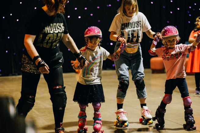 Two adults and two children wearing helmets and rollerkstaing in a hall against starlit backdrop
