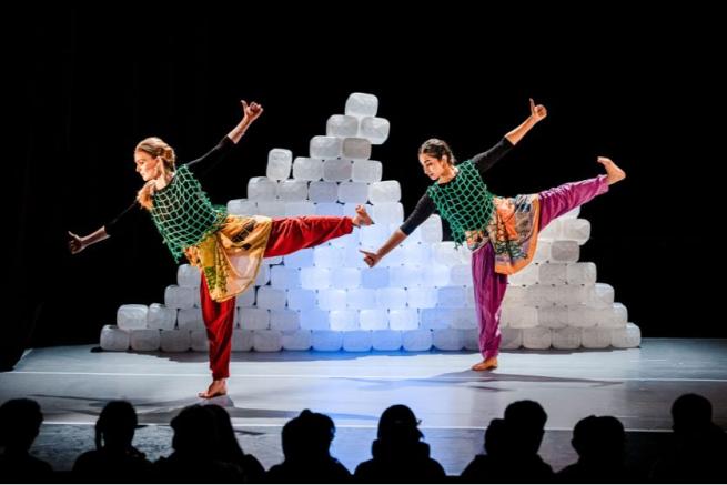 Two figures dancing on stage with a mountain of clear plastic tubs behind them. 