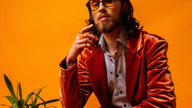 studio photo of Cezza. The image is very warm, in background and clothing with the jacket and glasses. Cezza looks at the camera very casually, but posed as such. A small green plant stands out, jutting out the bottom left corner of the image