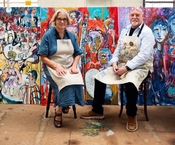Photo shows two Artists, Mel Davies and Kim Coley, Wearing paint  stained aprons, seated in front of large scale and colourful art works.