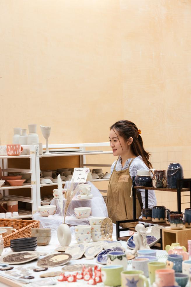 A woman in an apron stands at a pottery stall with handmade mugs, bowls, and jugs on display.