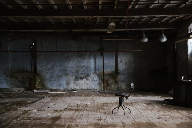 A dark lit old industrial mill room with a light blue peeling painted wall and uneven wooden flooring cover in dust and dirt. There are light and pipes hanging down and a single metal chair off to the right side.