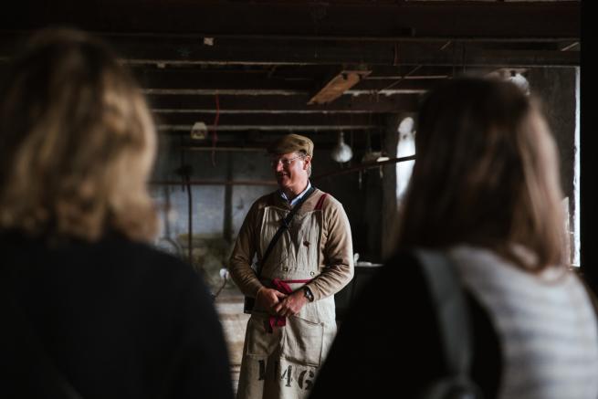 Managing Director William Gaunt stands within an old and dirty industrial room wearing a lined and red apron and flat cap. He is giving a tour and two people are seen beside him.