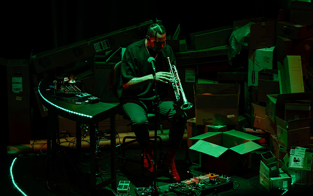 A man playing a trumpet sits beside a mixing desk, surrounded by cardboard boxes