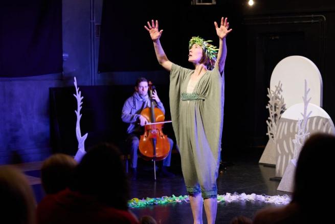 A dancing figure and cellist on performing on stage surrounded by cardboard cut-out trees. 