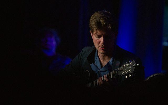 Tom Ollendorff playing guitar on a darkly lit stage