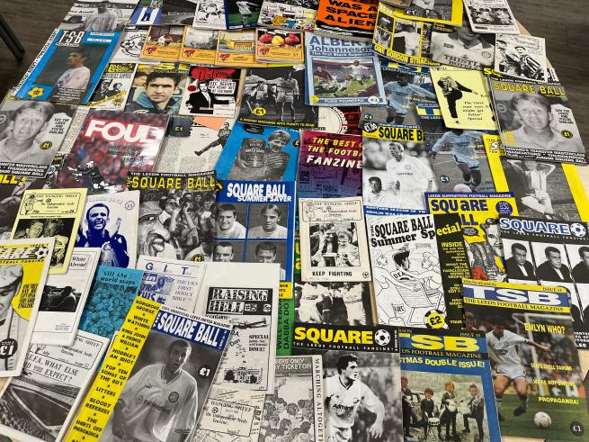 A wide assortment of football fanzines spread across a table, with many issues of "The Square Ball" visible. Covers vary in design, featuring football players, illustrations, and bold text in different colours. The fanzines overlap slightly, filling the entire tabletop.