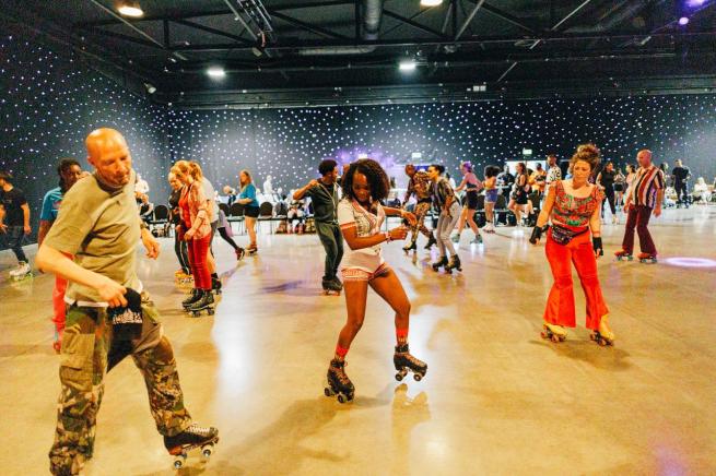 Adults roller dancing in hall against a starlight backdrop in bright vibrant clothing. 