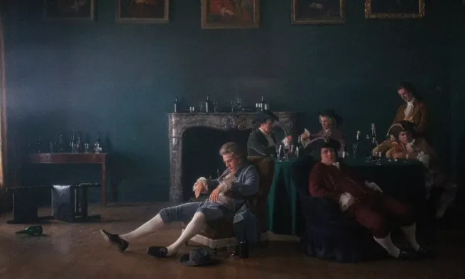 Barry slouches in an armchair while other men sit at a table behind him.