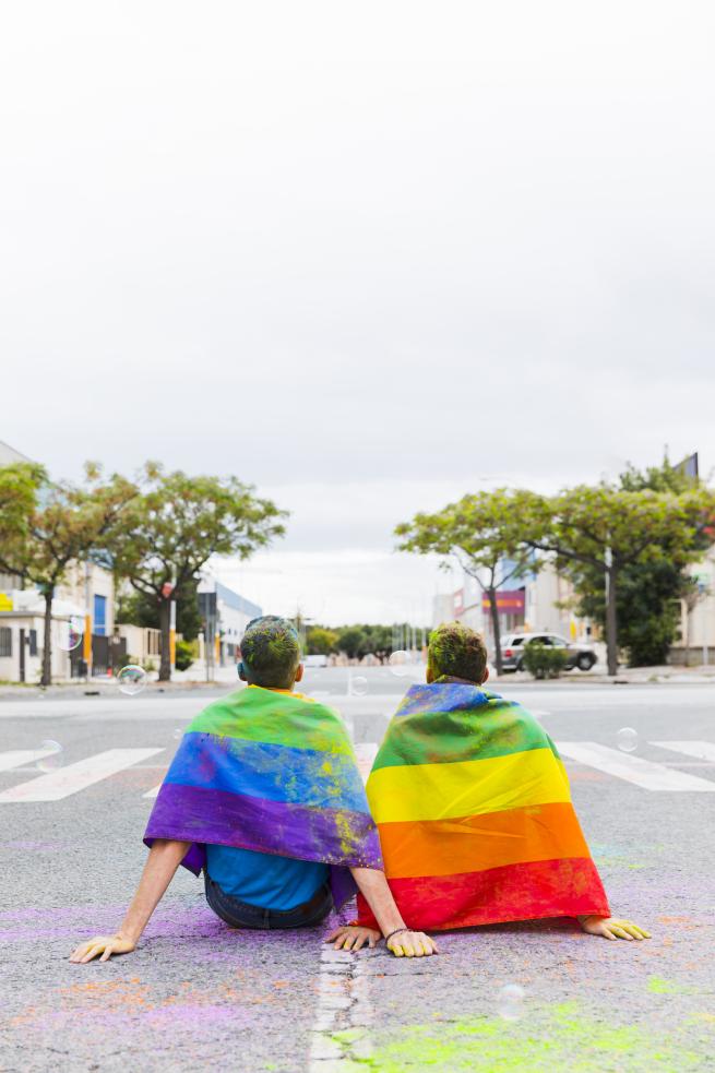 A picture of two people sat on a road wearing LGBTQ+ flags
