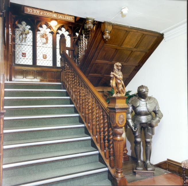 A picture of a Victorian staircase with modern stair treads, and a suit of armour at the foot of it. Faux-medieval stained glass windows are on the half landing.