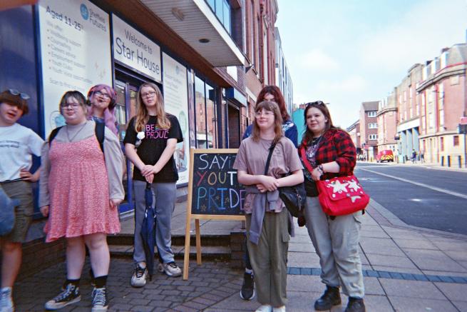 A group of young people dressed in colour clothes stood outside next to a sign that reads ''sayit youth pride''. They are wearing colourful clothes and smiling. 