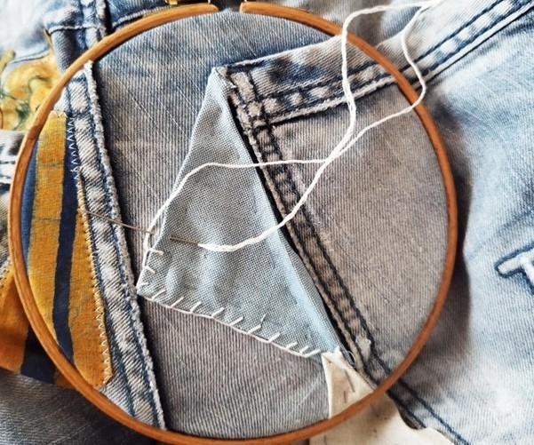 A wooden embroidery hoop on a pair of jeans with blue fabric being stitched onto it with white thread