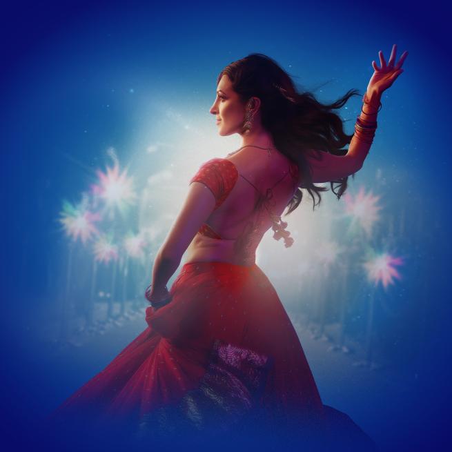 A Bollywood dancer posing while wearing a traditional red outfit with long flowing brown hair. Paparazzi cameras flash around her. 