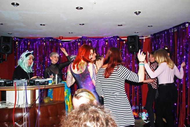 A group of girls dance on a dancefloor with purple glittery curtains. 
