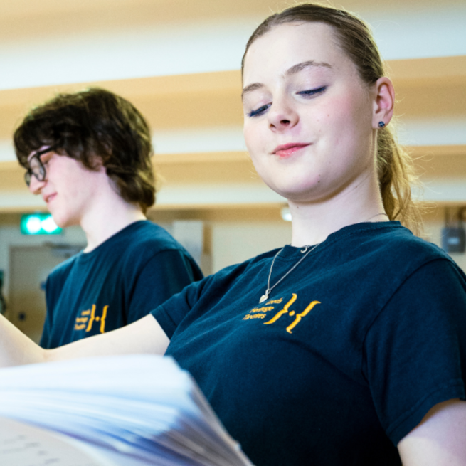 Young people are rehearsing and reading a script. They are wearing shirts with the Leeds Heritage Theatres logo.