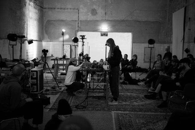 A black-and-white photo of two people operating music equipment in a dim, industrial-style room, while seated and standing audience members watch closely, surrounded by cables and lights.