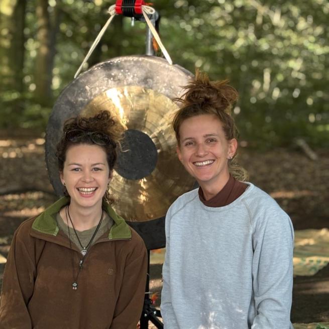 Two women smiling in a forest in front of a gong