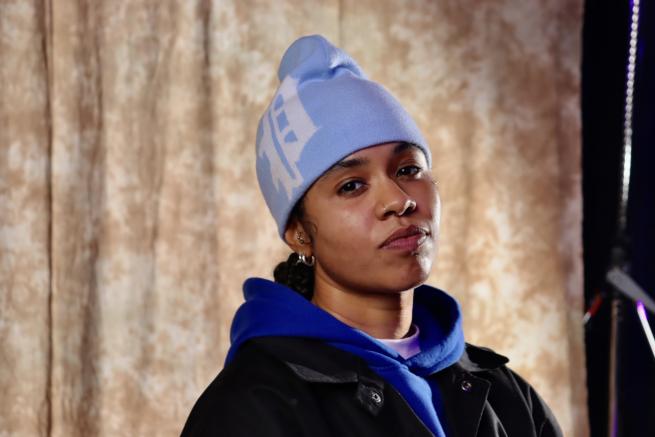 Che3kz and black female rapper in her 20s is wearing a light blue hat, a dark blue hoodie and a black jacket.t.