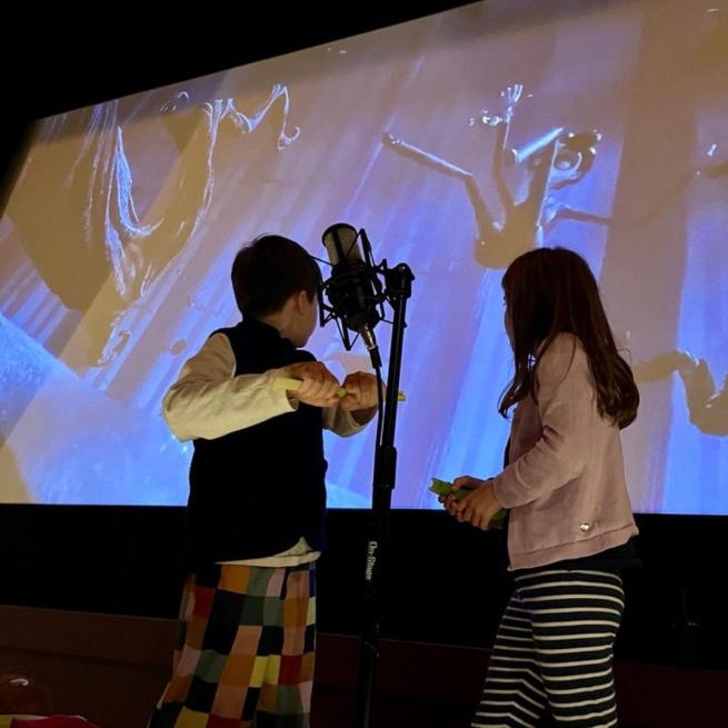 Two young people create sounds with props beside a microphone and large projection screen.