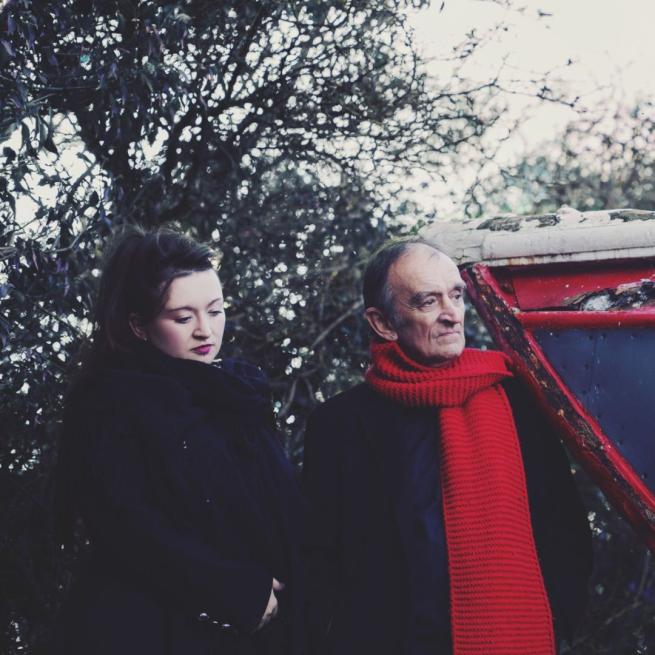 Martin and Eliza Carthy stand together in thick winter coats and long scarfs, both looking into the distance. The background is an icy scene with wintry trees and a dilapidated boat.