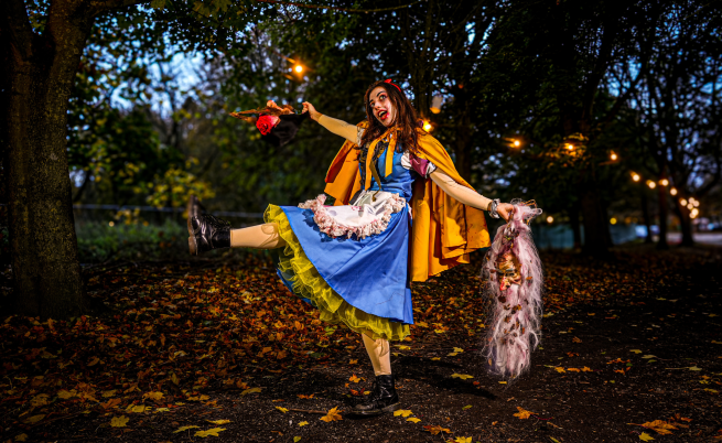 A dark woodland scene with a twisted version of Snow White standing with one leg out stretched and arms wide swinging halloween props