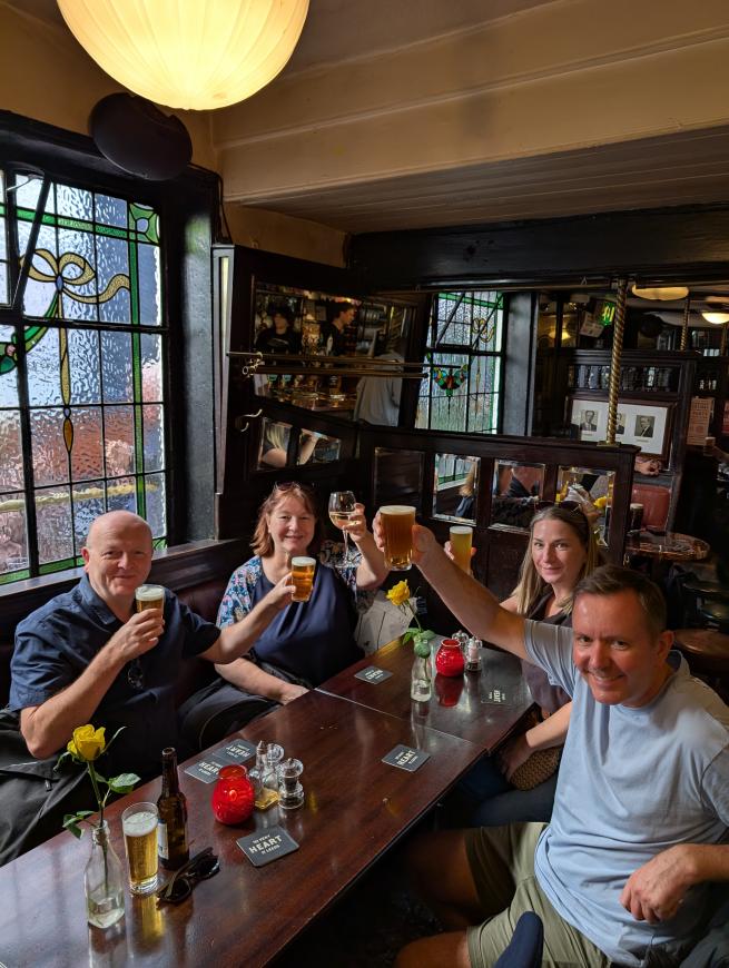 A group of four people inside a pub raising glasses of beer in toast towards the camera. The pub is Victorian-era decor.