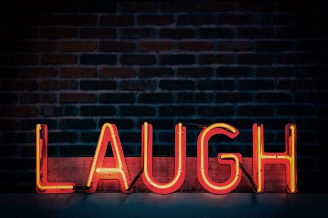 Photograph of the word Laugh in red neon letters against a dark brick wall.